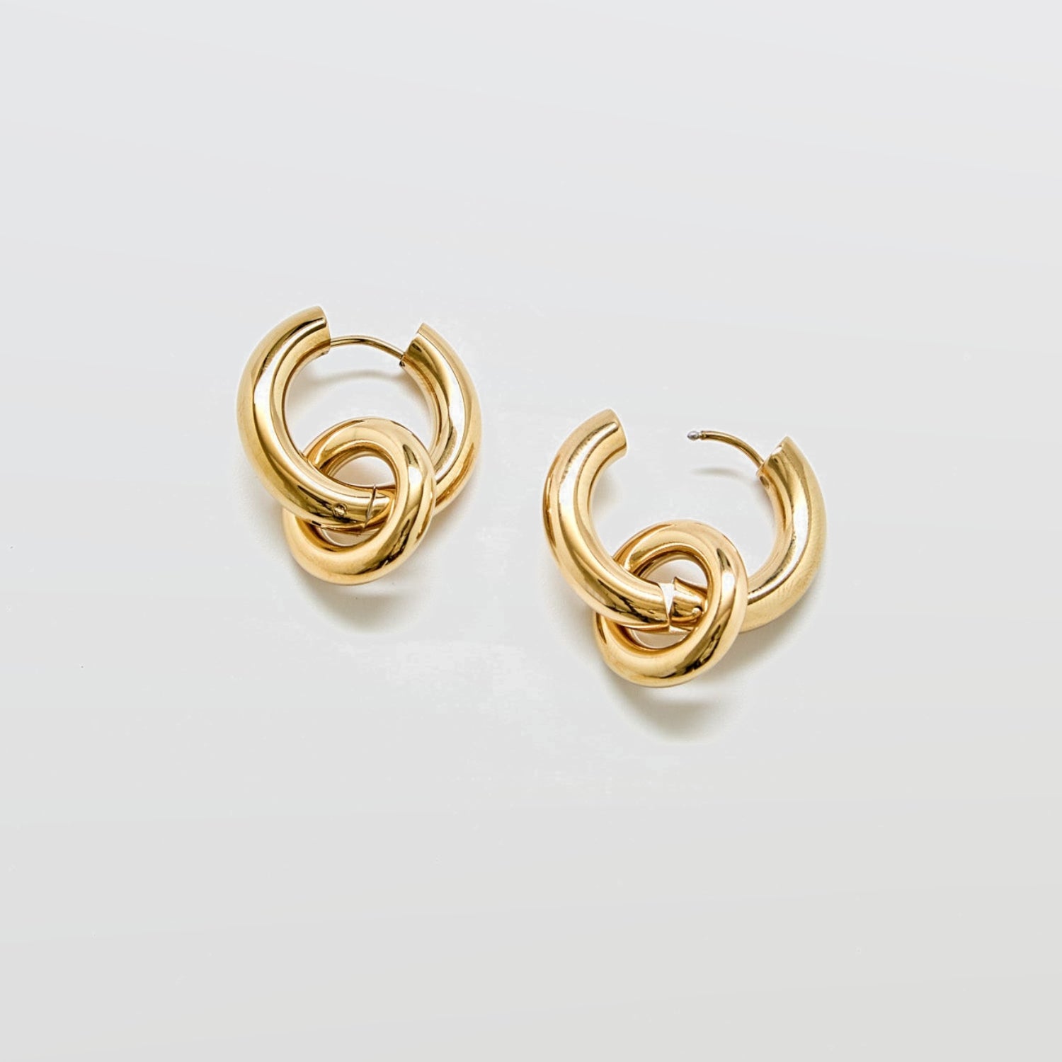 Polished gold stainless steel hoop earrings against white background featuring detachable interlocking loop charm, modern sculptural jewelry design.