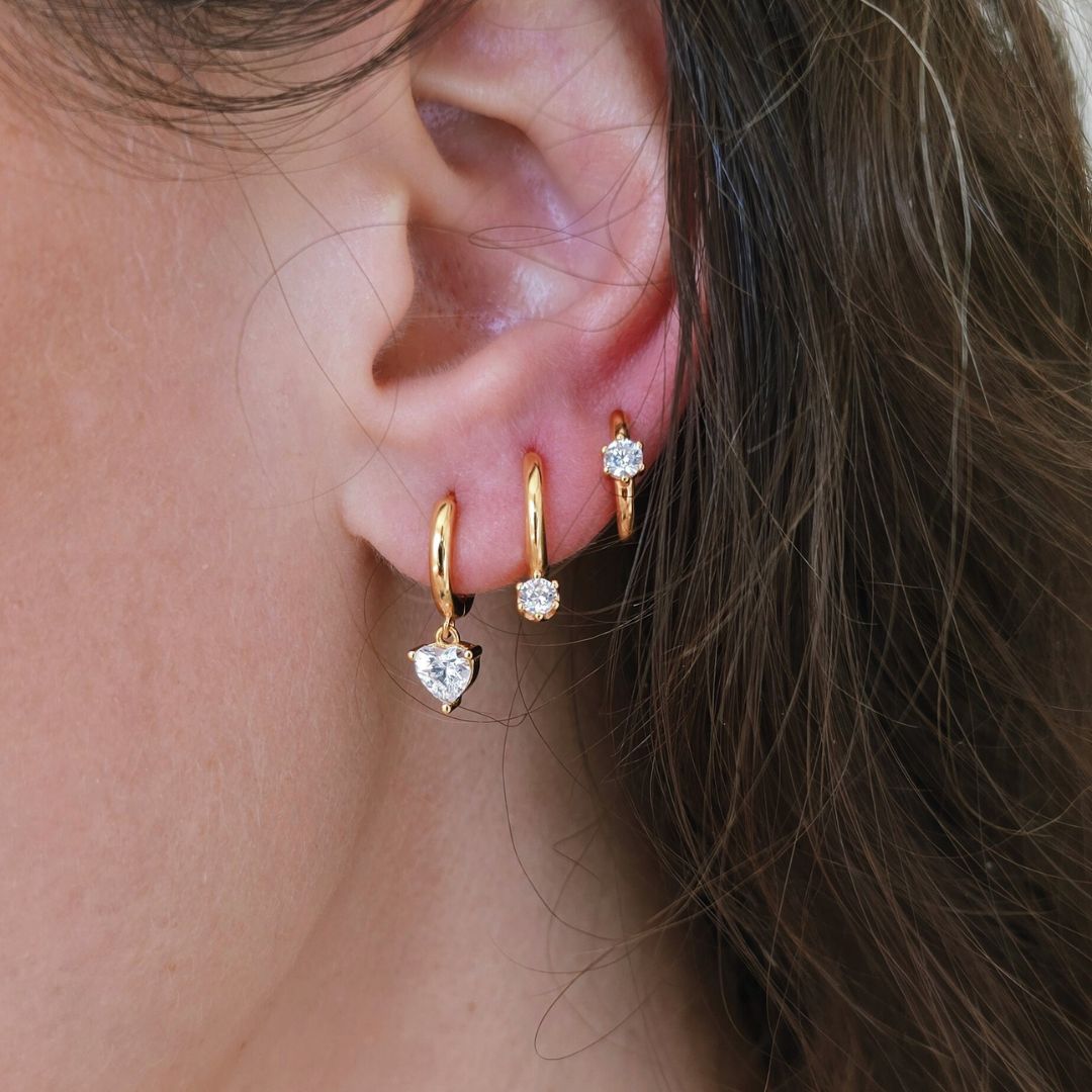 Gold CZ heart huggie earrings worn in an ear stack with gold cubic zirconia huggie hoops on ear.